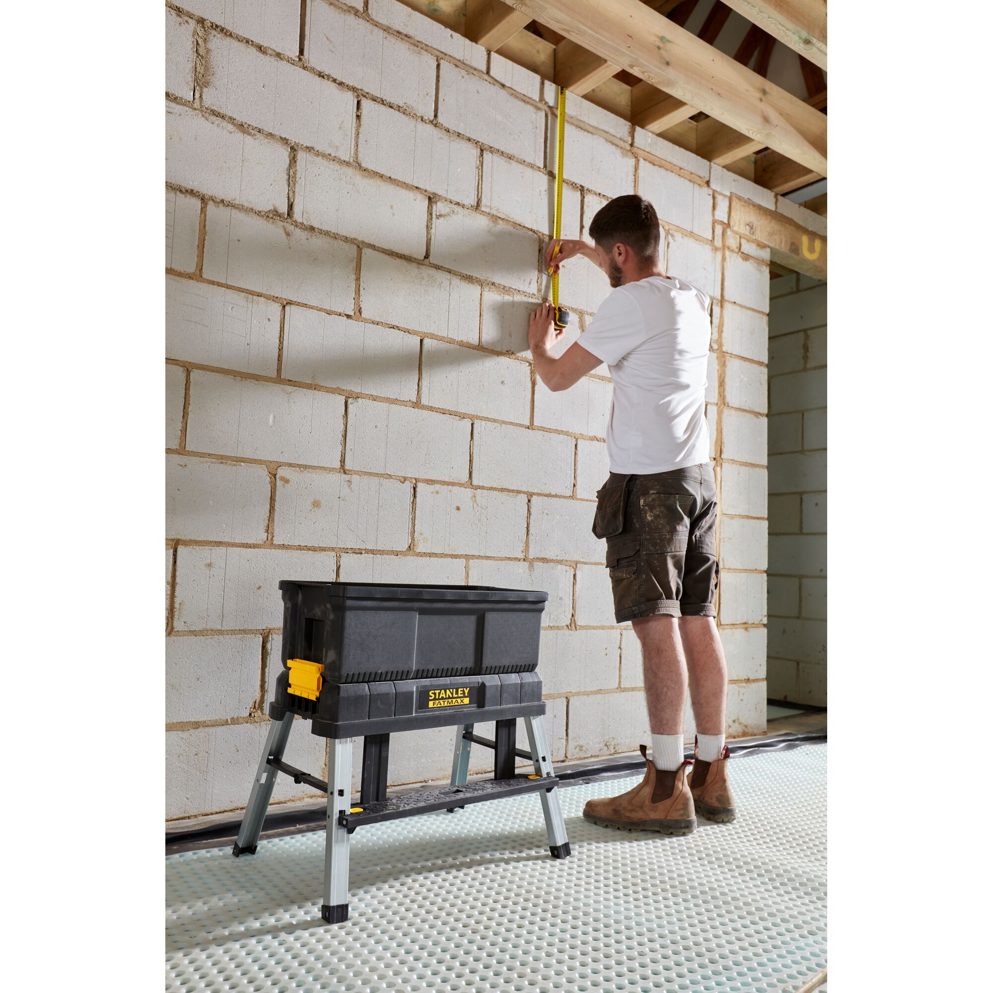 STANLEY FMST81083-1 black foldable sawhorse tool box on site, construction worker measuring wall.