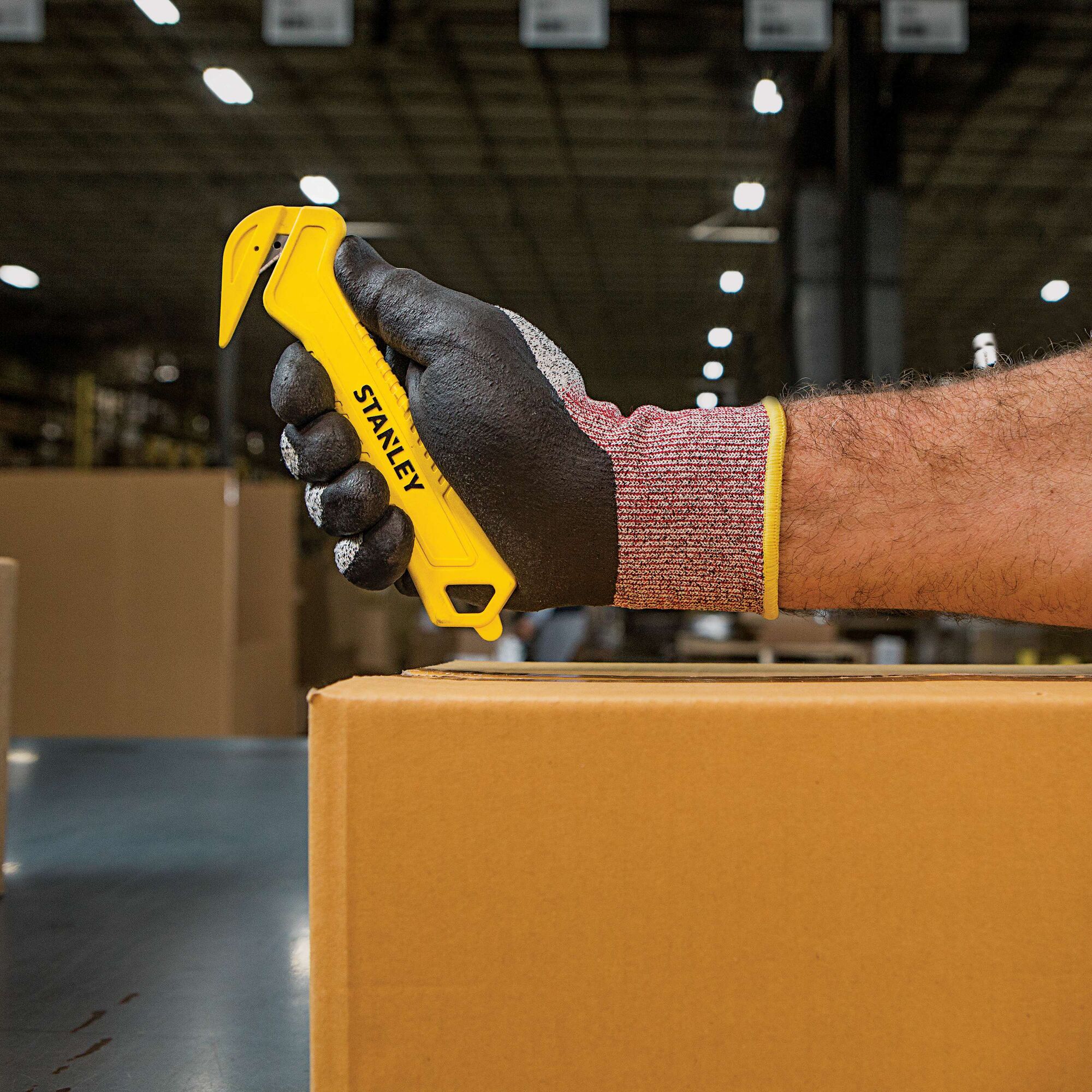 STANLEY STHT10355 yellow safety box cutter held by gloved hand cutting cardboard, warehouse background.