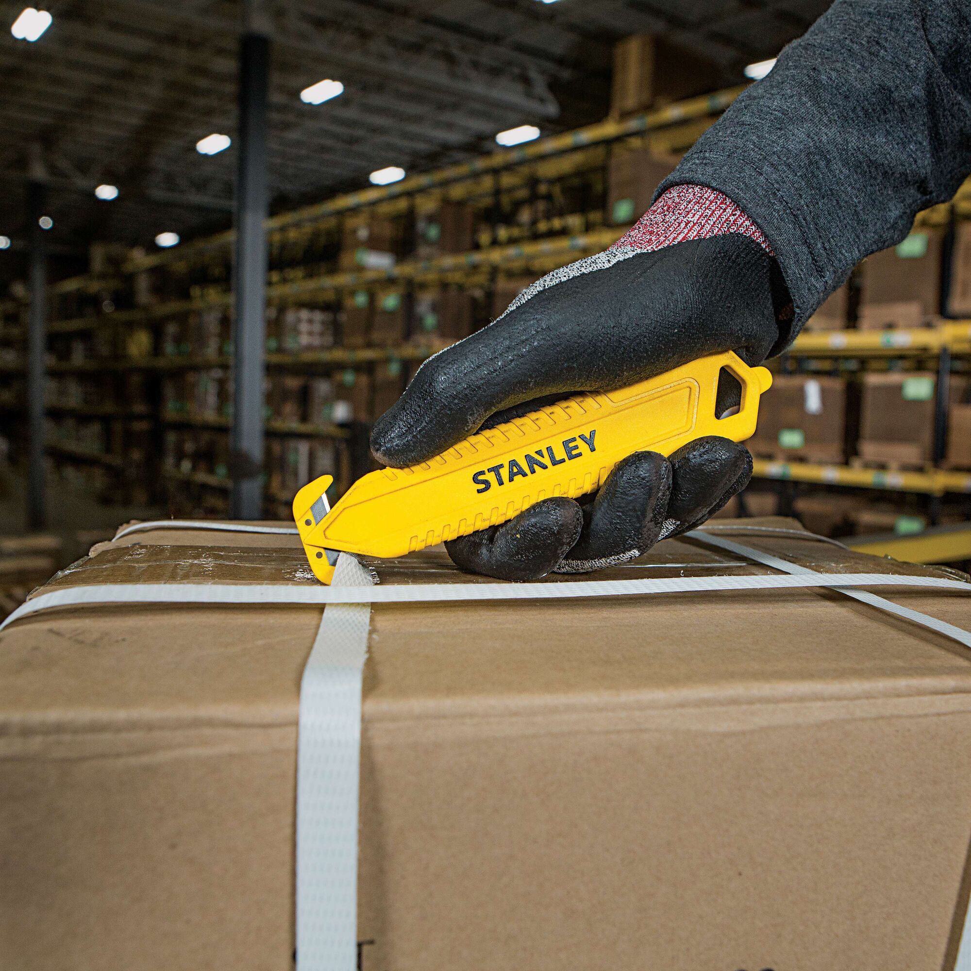 Stanley STHT10359 safety knife used to cut box straps, warehouse background, product held by gloved hand.