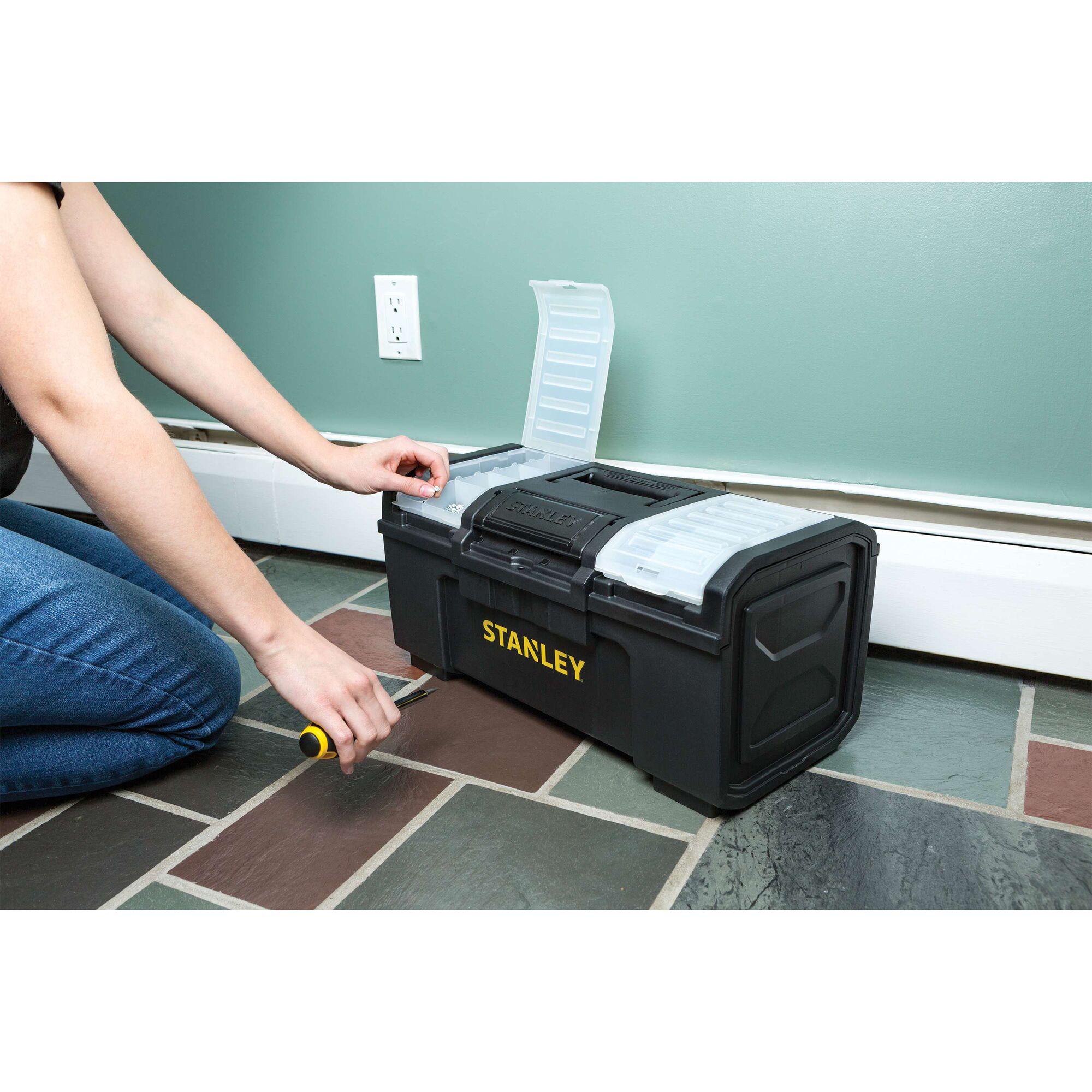 STANLEY STST19420 black plastic toolbox with open compartments, on floor, person using hand tool nearby.